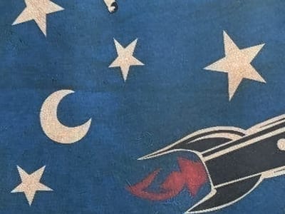 The image displays a close-up of a blue fabric with a pattern of stars, a crescent moon, and a rocket with red flames. The stars are light-colored and have five points. The crescent moon is also light-colored and positioned in the upper left quadrant of the image. The rocket is dark-colored with white accents and is depicted with red flames trailing from its rear, suggesting motion. The fabric appears to have a textured surface. The image does not provide any information about the city of Jrarat, Armenia, or any elements that would indicate a specific geographical location.