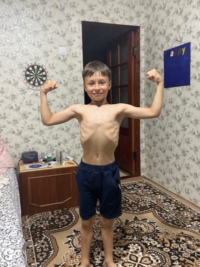 The media file shows a young boy in what appears to be a bedroom. The boy is shirtless and wearing dark blue shorts, flexing his biceps. He has short brown hair and is smiling at the camera. Behind him, a dartboard is hanging on the wall, and a patterned rug covers part of the floor. A wooden door is visible in the background, and the walls are covered in floral wallpaper. There's also a blue sign with "Happy" written in colorful letters hanging from the ceiling. On a table to the boy's right, there are several bottles and other items. The lighting suggests it might be indoors during the day or evening. The scene conveys a sense of casual playfulness.