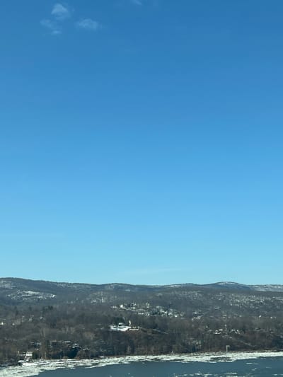 An elevated long shot captures a winter landscape in the Town of Cortlandt, United States of America. The scene is dominated by a clear, bright blue sky in the upper two-thirds, with a few faint white cirrus clouds visible in the upper left. A distant line of hills, partially covered in snow and bare deciduous trees, extends across the mid-ground. Scattered residential buildings with dark roofs are interspersed among the trees on the hillsides. The lower portion of the image displays a wide body of water, likely a river, covered with numerous irregular white ice floes. No individuals, explicit actions, or interactions are visible.