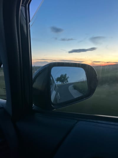 The view from inside a car looking out at the side mirror. The side mirror reflects a road, a tree and another vehicle. The sky and a strip of sunset are visible through the car window. The car's interior is visible, including the door frame and a portion of the dashboard. The location is unknown and there is no evidence to determine that it is in Blatnitsa, Bulgaria. No people are visible.