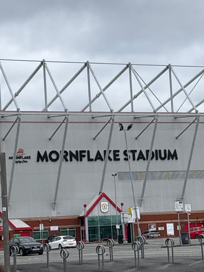 The image shows a view of Mornflake Stadium, located in Crewe, United Kingdom. The stadium facade is a large, ribbed white structure with the words "MORNFLAKE STADIUM" prominently displayed in black lettering. To the left of the stadium, there is a logo featuring a red crest with a bull. Below the logo, smaller text reads "Mornflake - Mighty Oats". The stadium is supported by a complex network of grey metal struts and beams, suggesting a large, modern construction.
In the foreground, there are several cars parked in what appears to be a parking lot or street. A black Audi is on the left, followed by a white SUV, and partially visible grey and red cars further down. Bicycle racks are also visible in the foreground, indicating public access. The sky is overcast, suggesting a cloudy day. The architectural style of the lower part of the stadium, with its red rooflines and glass entrances, suggests a fan zone or hospitality area.