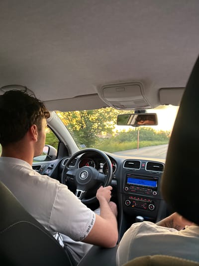 In Balanovo, Bulgaria, two individuals are captured from inside a car during a drive. The person in the driver's seat, a male with curly dark hair, is wearing a white t-shirt and is actively steering the vehicle. The car's interior is visible, showcasing the dashboard with a Volkswagen logo on the steering wheel, air vents, and a car stereo. The passenger, whose back is to the camera, wears a white t-shirt and has visible arm tattoos. The background through the car's windows reveals a rural landscape with trees bathed in the warm light of the setting sun, and a road stretching ahead. A rearview mirror reflects the driver's face and the surrounding scenery. The lighting suggests it is late afternoon or early evening.