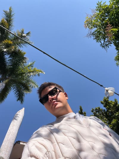 A low-angle shot frames a person with short hair and sunglasses, wearing a white cable-knit sweater. Overhead, a clear blue sky stretches behind palm trees and other green foliage. A light string with a bulb hangs diagonally across the frame. A white, cylindrical object appears at the bottom right, possibly a pillar or architectural detail. The perspective gives a sense of looking up at the subject and the surrounding environment.