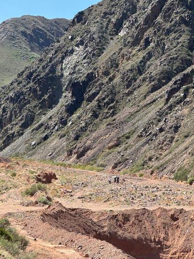 The image shows a steep, rocky mountain landscape in Krasnyy Most, Kyrgyzstan. The left side of the image consists of a dark, rugged mountain slope with sparse vegetation. The right side shows a lighter-colored, less steep slope with a walking path. Three people are standing together on the path. The sky is visible as a sliver of blue in the bottom left corner.