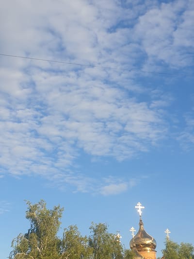 The image shows a bright blue sky with scattered white clouds. In the lower portion of the image, the tops of several trees with green foliage are visible, along with the golden dome of a church topped with crosses. A single black wire stretches horizontally across the upper part of the sky. The scene is bright and appears to be outdoors during daylight hours. No people are visible in the frame.