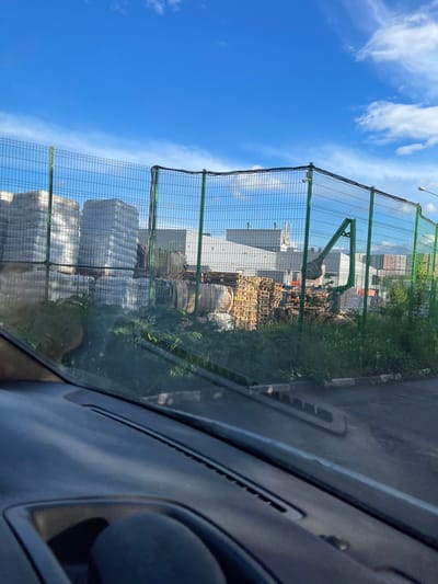 A view from inside a vehicle shows a fenced-off industrial area in Bittsa, Russia. Visible through a green wire mesh fence are stacks of white-wrapped materials, piles of wooden pallets, and industrial buildings. The sky above is blue with scattered clouds. Overgrown grass grows along the base of the fence. A portion of the vehicle's dashboard and window frame is visible on the right side of the image.