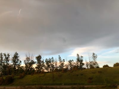 This is a landscape photograph taken from a moving vehicle, likely a car or train, as indicated by the motion blur. The scene depicts a grassy hill lined with trees, set against a cloudy sky. The time of day appears to be late afternoon or early evening, with the setting sun casting a warm glow on the landscape, visible as hints of light breaking through the clouds and illuminating the grass. The weather is overcast, with large, dark clouds dominating the upper portion of the frame, suggesting the possibility of rain or a coming storm. The trees are a mix of deciduous, with some bare branches, and evergreens, indicating an autumn setting. The location is Innisfil, Canada. There is no visible text in the image. The overall mood is somewhat somber due to the heavy clouds, but the warm light on the hill offers a touch of hope.