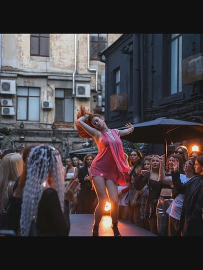 A young woman with red hair and a short pink dress is dancing on a raised platform in an outdoor urban setting. She is captured mid-motion, with her arms outstretched and her body curved. A crowd of people, mostly women, watches her from below. The background features a building with multiple windows and air conditioning units, suggesting a residential or commercial area. The scene is lit by artificial lights, creating a dramatic effect. The exact location in the city of Norcia, Italy is not discernible from the image.