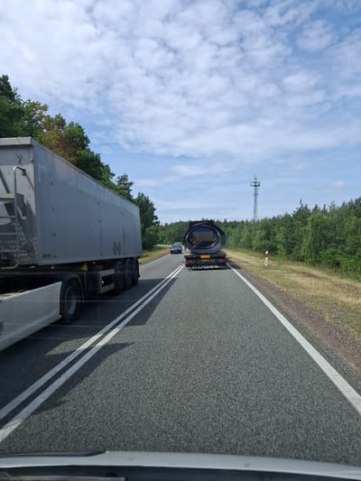 The image depicts a two-lane asphalt road bordered by a grassy shoulder and a dense forest. A large semi-trailer truck with a grey cargo box is in the left lane, its front and left side visible. In the right lane, another semi-trailer truck is ahead, carrying a large, dark, circular object on its trailer. A small, dark-colored car is visible in the distance, also in the right lane. A cell tower stands tall in the background, situated beyond the treeline. The sky is partly cloudy with scattered white clouds against a blue backdrop. The scene appears to be a daytime shot on a highway or a major road in Przyłubie, Poland, given the context.