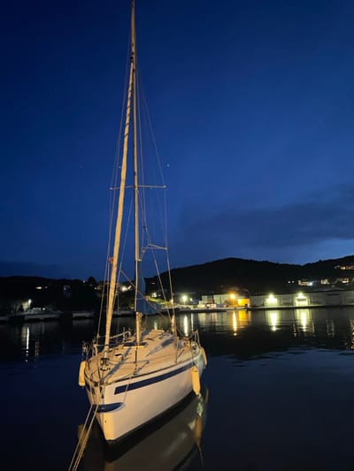 A sailboat is docked at night, its mast towering against a dark blue sky. The boat is white with a dark stripe, and its sails are furled. Dock lights reflect on the calm water. A dark, tree-covered hill rises in the background. Buildings are visible along the shoreline. The scene exudes a peaceful, nautical atmosphere.
