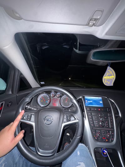 The image captures the interior of a car, likely an Opel, at night or in very low light, set in Musachevo, Bulgaria. A person's right hand, wearing blue ripped jeans, rests on the multi-function steering wheel. The car's instrument cluster shows a speed of 0 km/h, a trip meter reading "238.9 km", and a total odometer reading of "252910 km". Notably, an engine warning light is illuminated.

The dashboard features an infotainment system displaying "Audio FM" and "108.0 MHz," along with a temperature reading of "6°C". Below the screen are various climate control buttons, including "AUTO," and other controls for media and phone functions. A blue illuminated car charger is visible in the console to the right. Hanging from the rearview mirror is a yellow air freshener with the text "areon mon areon". The exterior view through the windows is almost completely dark, reflecting interior lights. The car's interior trim is light gray on the pillars and roof, contrasting with the dark dashboard and black leather steering wheel.