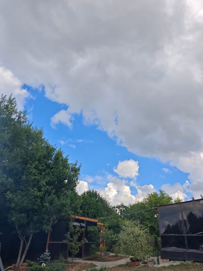 The image depicts an outdoor scene in the village of Лужники Снт, Russia. The sky is partially cloudy with patches of bright blue sky visible. Lush green trees line the foreground and midground, with a variety of deciduous and evergreen species. Several structures are present, including what appears to be a covered patio or gazebo with a dark, translucent covering on the side, and another partially visible structure to the left with an orange-tinted frame. String lights are visible hanging between trees. The ground is a mixture of dirt, gravel, and some landscaping elements. No people are visible in the image. The overall impression is that of a backyard or outdoor recreational area on a day with variable weather conditions.