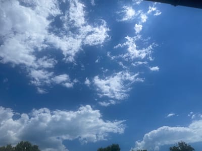 The image shows a bright blue sky with scattered white cumulus clouds. The sun is partially visible, casting bright light in the upper right portion of the frame. A dark, curved object, possibly a roof overhang or gutter, is visible along the very top edge of the image. The lower edge of the image shows the tops of several green trees, indicating an outdoor, natural setting. The scene appears to be a clear day. The location is specified as Ливадийский поселковый совет, Ukraine.