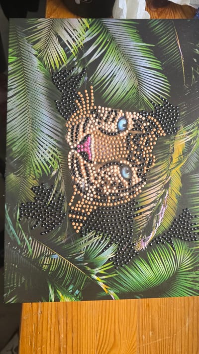 The image shows a diamond art depiction of a leopard's face emerging from a background of lush green palm leaves. The leopard's face is composed of small, round beads in shades of tan, black, and pink, with bright blue eyes. The background is a dark green with lighter green palm leaves. The entire image is on a rectangular black surface, possibly a canvas or board, and sits on a wooden table with a yellow finish. The location (Zirndorf, Germany) is unknown and cannot be determined from the image.