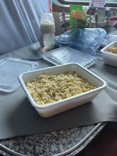 A close-up shot shows a white plastic container filled with instant noodles on a gray surface, possibly a table in a restaurant or a train. To the left, a partially visible white plastic lid with condensation sits on the surface. In the background, a baby bottle with a white nipple and clear liquid is visible, along with various containers, cups, and a green packet of what appears to be seasoning. The overall scene suggests a casual meal setting. There is no information to suggest this scene is in Kizner, Russia.