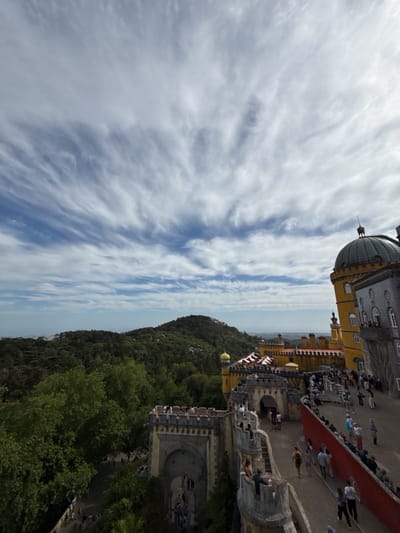 The image depicts a high-angle view of the Pena Palace in Sintra, Portugal. A sprawling castle complex dominates the right side, featuring yellow and gray towers, domes, and elaborate architectural details. Numerous people are visible along the pathways and walls of the palace, indicating tourist activity. A red wall borders a walkway populated with visitors. In the background, a densely forested hillside stretches toward the horizon. The sky is partially cloudy, with wispy clouds spread across a predominantly blue expanse. The overall scene captures the grandeur of the palace and its setting amidst lush greenery.
