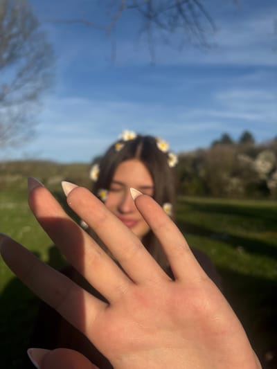 A close-up, slightly out-of-focus photograph shows a fair-skinned hand with long, pointed nails, positioned in front of a partially visible person. The hand is in focus, and light casts shadows across the fingers. The person in the background has brown hair adorned with small white daisy-like flowers. The backdrop consists of a blue sky and green foliage, indicating an outdoor setting. Based on the presence of light and vegetation, it appears to be a park or garden. While the location is specified as Beli Brod, Bulgaria, there is no information in the image that allows us to infer this beyond the geographical data provided in the prompt. The image is oriented sideways with the sky on the left and the ground on the right.