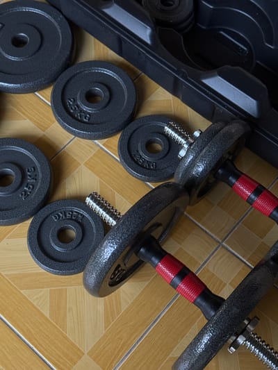 In Kalirancang, Indonesia, an overhead shot shows a set of dumbbells and weight plates arranged on a tiled floor. The dumbbells have black textured grips with red accents. Several round, black weight plates are scattered around the dumbbells, with some bearing the inscription "2.5 KG". A black plastic carrying case for the dumbbells is partially visible in the upper right corner of the image. The tiled floor has a pattern of light brown and beige squares.
