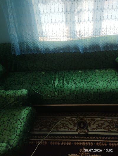 A green sofa with a patterned cover is in the foreground. A blue lace curtain hangs in front of a window behind the sofa. A carpet is on the floor in front of the sofa. A white cable is visible on the carpet. The date stamp "30.07.2026 13:02" is in the lower right corner of the image. The scene is indoors, likely a living room in a residence in the city of Quyosh, Uzbekistan.