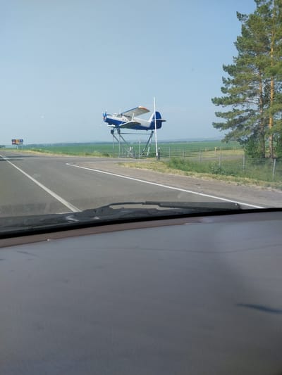 A blue and white biplane is mounted on a metal stand next to a road in Belokurikha, Russia. The plane is positioned as if it is taking off, with its nose pointed upward. The road appears to be a highway, with white lines marking the lanes. In the background, there are green fields and a large pine tree. The sky is clear and blue. The image is taken from inside a vehicle, looking out through the windshield. A dashboard is visible in the foreground.