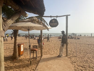 The image shows a beach scene in Saly Portudal, Senegal, at Safari Beach. A crowd of people is visible on the beach and in the ocean. A wooden sign reads "Safari Beach" above the entrance to the beach. A man wearing a black shirt with "security" printed on the right shoulder and light-colored pants leans against a wooden post. A thatch-roof structure, an umbrella, a small table with a chair, and a trash can are visible on the left. The environment is sandy with a clear, light-colored sky.