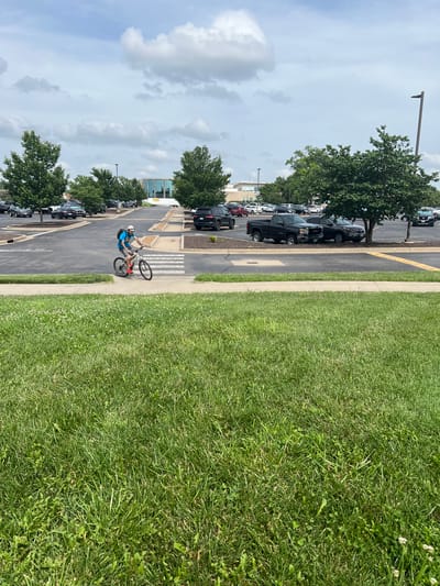 A person rides a bicycle on a crosswalk at a parking lot in Columbia, MO. The person is wearing a blue shirt, grey shorts, a white hat, and a backpack. The bicycle is black. The parking lot has several parked cars and trees. The sky is cloudy. The foreground is a grassy lawn.