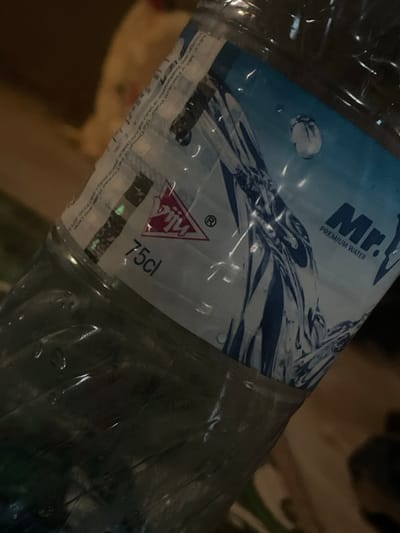 The image depicts a clear plastic water bottle, positioned diagonally and captured in a dim, indoor setting in Mara, Nigeria. The bottle features a label that identifies the product as "Mr. PREMIUM WATER" with a blue and white design showing water splashes. A red triangular logo with "Piu" and an ® symbol is visible, along with the volume "75cl" printed in black. Partially legible text on the label's left edge includes "MKV INDUSTRY" and a copyright date of "2019." The bottle's exterior exhibits multiple water droplets or condensation. The background is out of focus, showing indistinct shapes in shades of brown and beige, with a lighter object visible in the upper left quadrant. No individuals or active interactions are visible in the scene.