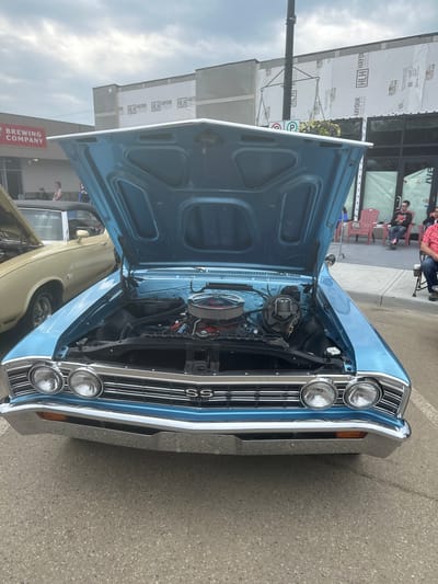This media file captures a close-up, high-angle shot of the front of a light blue classic car, likely a Chevrolet Chevelle SS, with its hood open. The car is parked on a paved surface, possibly a street or parking lot, with a building and some people visible in the background.
The car's engine is exposed, revealing a chrome air cleaner and various engine components. The front grille, headlights, and chrome bumper are highly polished. The "SS" emblem is clearly visible on the grille. To the left of the classic car, the front of a lighter-colored vintage car is also partially visible.
In the background, a building with large windows is under construction, covered in white sheeting that displays "HLH HAYDUK LUMBER AND HARDWARE" and "TITAN" text. A red sign with white lettering reads "BREWING COMPANY". Several people are seen standing and sitting in the background, some on chairs, suggesting a car show or community event. The lighting suggests it is daytime, with overcast or partly cloudy skies. The overall impression is of a car enthusiast gathering or a local event showcasing vintage automobiles.
