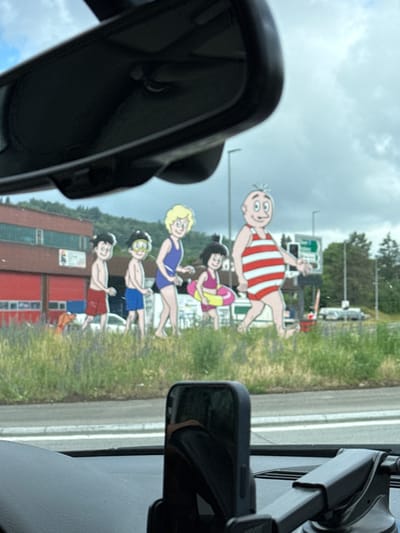 The image displays a series of five cheerful, flat cartoon cutouts depicting a family in swimwear, positioned alongside a road. The scene is viewed from inside a car, with a rearview mirror visible on the left and a device mount on the right.

The largest cutout, in the foreground, is a man with a large belly wearing a red and white striped one-piece swimsuit, smiling broadly with spiky hair. Behind him are four smaller figures: a woman with dark hair in a purple swimsuit holding a yellow inflatable ring, followed by a blonde woman in a blue swimsuit. Next is a boy with dark hair, a yellow swim mask, and blue swim shorts, and finally, another boy with dark hair and red swim shorts. All figures have happy, smiling expressions, suggesting excitement for a swimming or water-related activity.

The background features a grassy verge, leading to a tree-lined area and distant buildings, including a prominent red structure and a white one. The sky is overcast with visible clouds, indicating daytime. The cutouts appear to be a promotional display, likely for a public bath, thermal spa, or swimming facility, consistent with the location context of Zurzach, Switzerland, which is known for its thermal baths. The overall impression is a welcoming and fun atmosphere, indicative of leisure or vacation. No clear, readable text is visible on the cutouts or in the distant background elements.