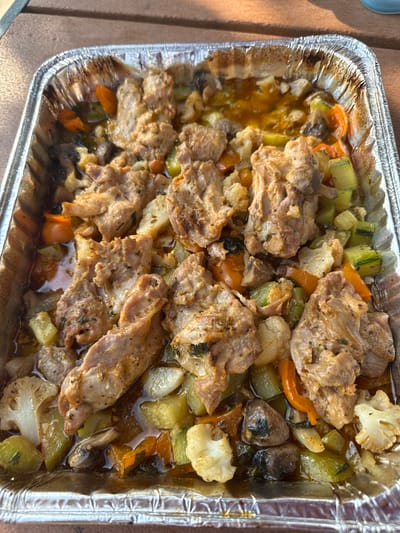 A foil pan filled with roasted meat and vegetables sits on a wooden table. The meat appears to be chicken or pork, cut into bite-sized pieces and seasoned. Among the vegetables are cauliflower florets, sliced zucchini, carrots, and mushrooms. The dish is coated in a light sauce or juices, indicating it was likely baked or roasted. The lighting suggests it is daytime, with sunlight casting shadows. The background is blurred but appears to be an outdoor setting, possibly a backyard or patio. The image was taken in Allen Township, United States of America.