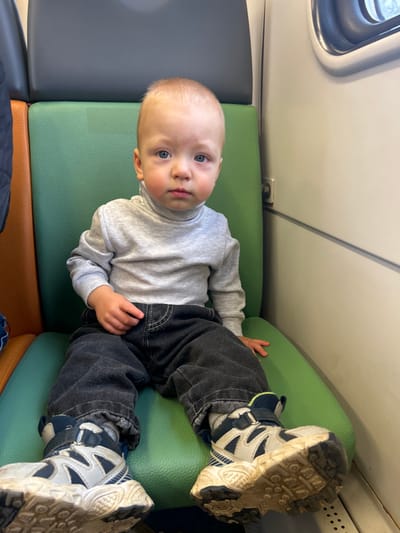 The image depicts a young child seated on a passenger vehicle, presumably a train or bus, in the city of Peski, Russia. The child, with light-colored hair and blue eyes, appears to be a toddler and is dressed in a light grey ribbed turtleneck long-sleeve shirt, dark grey loose-fitting pants with white stitching details, and bulky white, blue, and black athletic shoes. The soles of the shoes are visibly soiled with dirt. The child is positioned centrally in the frame, facing directly towards the viewer with a neutral expression, and has noticeably reddish cheeks.

The child is seated on a bright green, textured, padded seat, which features a darker grey upper portion. To the left, a section of an adjacent seat, appearing orange-brown, is visible. The background on the right side of the image reveals a plain, light-colored wall with a prominent horizontal seam or stripe. A rounded rectangular window is visible in the upper right quadrant of the image, allowing external light to enter. A small, dark, circular object is affixed to the wall below the window, and a metal grate or vent is partially visible at the bottom right. The overall environment suggests an interior compartment of a public transport conveyance.