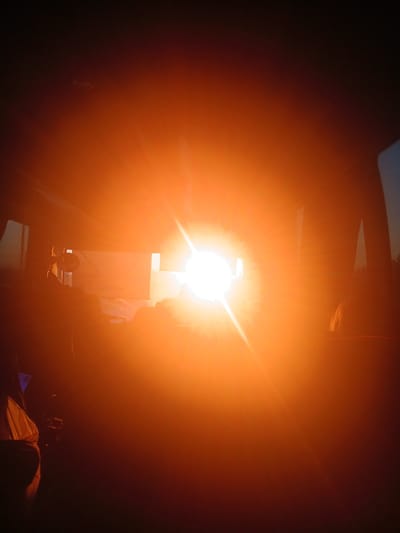 The image depicts an indoor scene dominated by a bright, concentrated light source. The light is orange-tinted and obscures much of the surroundings. Silhouetted forms of objects are visible in the foreground and midground. A tall rectangular shape can be discerned in the center, possibly a box or a screen, directly in front of the light. To the right of the light source, the vague outline of a person's head and shoulders is visible, also silhouetted. The background is dark and indistinct. No elements indicate that the location is Ustinovo, Russia.