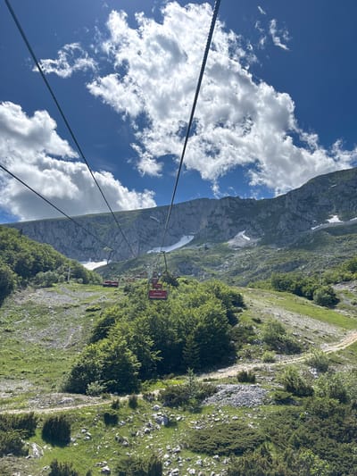 The image captures a scenic view of a ski lift traversing a mountainous landscape in Motički Gaj, Montenegro. The ski lift, with its red and yellow cabins, ascends towards a majestic, sheer rock face. The slopes are covered in lush green vegetation and patches of snow, indicating a transition between seasons. A dirt path winds its way up the hillside. The sky above is a vibrant blue, dotted with fluffy white clouds. The overall impression is one of natural beauty and recreational activity within a rugged mountainous environment.