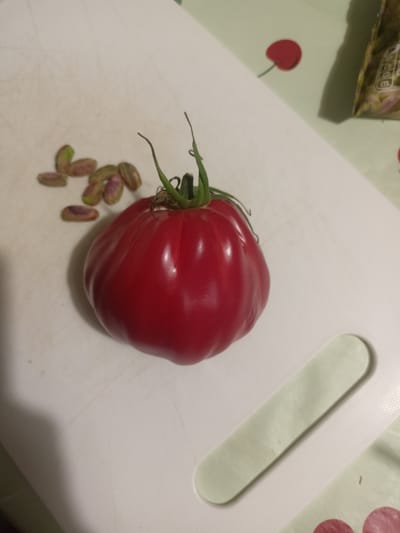 The image depicts a close-up, overhead view of a red tomato with a green stem and leaves, situated on a white cutting board. Scattered to the left of the tomato are several peeled pistachios, some showing pinkish hues. The cutting board has faint lines and a textured edge. In the background, out of focus, is a surface with a cherry pattern, and part of a package with text. This scene is on a white cutting board which is on a table in a kitchen. The location is not specified in the image or the provided context to be Grudziądz, Poland.