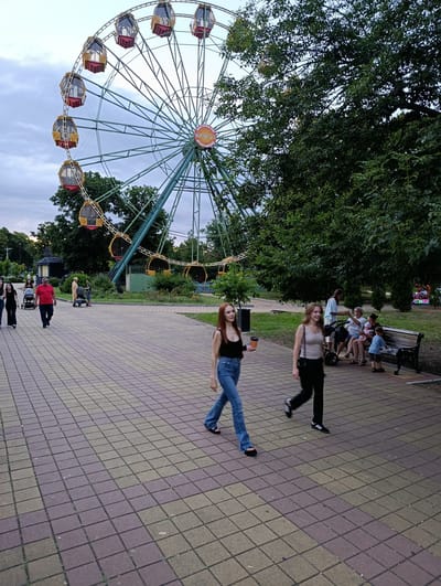 In Kelermesskaya, Russia, on a paved walkway with brown and beige square tiles, two young women are walking towards the viewer. The woman on the left has long, light brown hair and is wearing a black tank top and blue jeans. She is holding a cup in her right hand. The woman on the right has blonde hair and is wearing a beige tank top and black pants. She is holding a crossbody bag on her left side. In the background, to the left, is a large Ferris wheel with several gondolas, some of which are yellow and red. Trees with green foliage are visible behind the Ferris wheel and to the right of the scene. Further in the background, on the right, several people are sitting on a dark-colored park bench under the shade of a large tree. There are also strollers visible near the bench. The overall scene suggests an amusement park or a public park setting on a cloudy day.