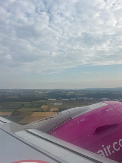 This media file, captured from an airplane window, shows a view of the landscape below and the wing and fuselage of the aircraft. The scene is oriented vertically in the image, suggesting the camera was held sideways or the photo was rotated.

The left side of the image is dominated by a vast expanse of clouds, ranging from fluffy white cumulus to more diffused, layered stratus clouds. The sky appears bright, indicating it is daytime, likely late morning or early afternoon. The weather seems partly cloudy.

The right side of the image reveals the wing and part of the fuselage of an aircraft. The fuselage is painted a vibrant pink and has white lettering that reads "air.co". Several warning labels are visible on the pink surface, including "NO STEP" and "NO GRAB". The wing is predominantly grey and white, with visible rivets and panel lines, typical of aircraft construction. Towards the bottom right, a circular insignia with red and white segments is partially visible on the aircraft.

Below the aircraft, a landscape unfolds, showing a patchwork of agricultural fields in shades of green and yellow, interspersed with darker patches of trees and a densely built-up area that appears to be a town or city. The overall impression is of an aerial view of a rural or semi-rural area.

The photograph appears to be taken while the aircraft is in flight, possibly during ascent or descent, given the angle and the close proximity to the landscape. There is no indication of people or any specific activity taking place beyond the act of flying. The location context provided is Echterdingen, Germany, which aligns with the visual cues of European-style agriculture and architecture.