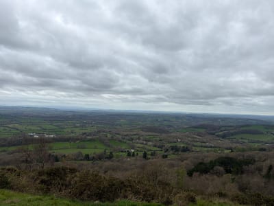 The image presents a vast landscape from an elevated vantage point in the Malvern Hills, United Kingdom. The scene is dominated by a cloudy, overcast sky. Below, a sprawling patchwork of green fields and wooded areas stretches into the distance. The landscape is punctuated by clusters of buildings, possibly farmhouses or small villages. The foreground shows vegetation, including scrubby bushes and grass, indicating the viewpoint's elevated position. There are no people visible in the frame. The overall impression is one of a rural, hilly landscape under a heavy, cloudy sky.