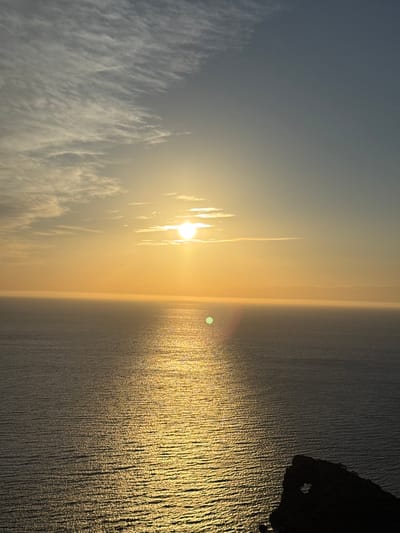 A photograph of the ocean horizon during sunset in Deià, Spain. The water is glistening gold from the sun. A rock is visible on the top right. Some clouds are to the left, and the sky is a grey-blue color. A slight lens flare of the sun is visible on the water.