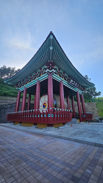 The image depicts a traditional Korean building with a distinctive green, multi-tiered roof and red cylindrical pillars. The building appears to be a pavilion or a gateway. A large drum is visible inside, adorned with a Taegeuk symbol, indicating a cultural or ceremonial purpose. The architecture features intricate details, including decorative carvings on the eaves and ornate brackets supporting the roof.

The ground in front of the structure is paved with interlocking tiles, forming a pathway. The surrounding environment includes a grassy slope with trees on the left and a stone wall on the right. The sky is visible above, with some wispy clouds, suggesting it is daytime, possibly morning or late afternoon due to the soft lighting.

There is visible text on a dark plaque attached to the building. The characters are Korean and appear to read "천고각" (Cheongogak). The overall scene evokes a sense of cultural heritage and tranquility. No people are visible in the image. The photograph is taken from a low angle, emphasizing the grandeur of the structure against the sky. The location is identified as Godang-ri, South Korea, reinforcing the Korean architectural context.