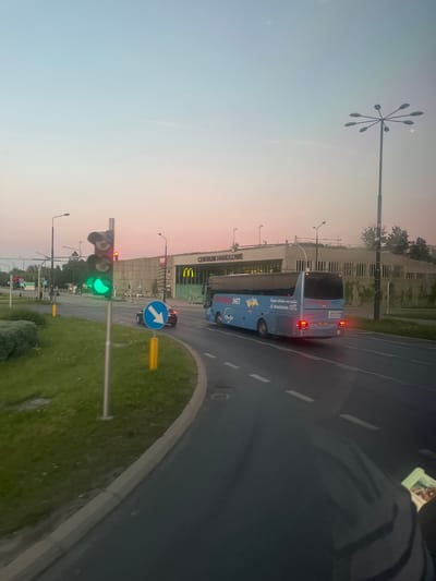 The image captures a street scene in Lublin, Poland, at dusk. In the foreground, a dark road with white lane markings curves to the left. A green traffic light is illuminated, indicating that traffic may proceed. A blue bus with "ORIONBUS.NET" branding is traveling on the road. In the background, a modern building with the text "CENTRUM HANDLOWE" (Shopping Center) and a McDonald's logo is visible. The sky displays a soft gradient of pink and blue hues. A tall street lamp with multiple fixtures stands to the right of the frame. The overall environment suggests an urban setting during a transitionary period of the day.