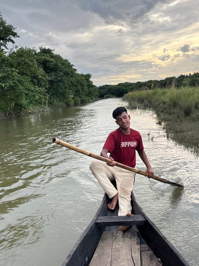 A young man in a maroon t-shirt with "Ferrari" printed on it and light-colored pants is sitting in a wooden boat on a river. He is holding a long wooden pole, appearing to be rowing the boat. The boat is moving through the water, which is reflecting the cloudy sky. Lush green trees and vegetation line the banks of the river, characteristic of the Sylhet region of Bangladesh, known for its natural beauty and waterways. The sky is overcast with dramatic clouds, suggesting either early morning or late afternoon lighting. The scene captures a moment of serene travel through a natural landscape.