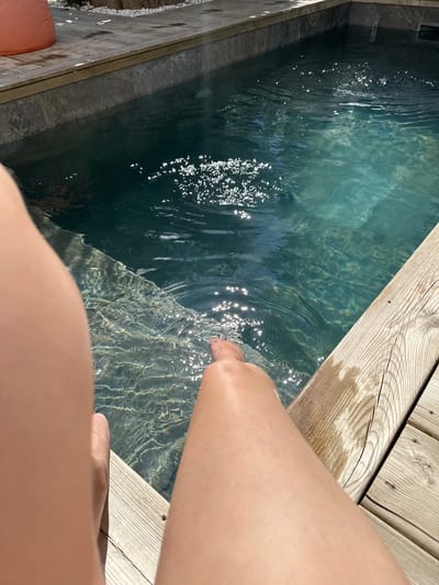 A medium, angled shot depicts a partially visible individual dipping their foot into a pool of water. The setting includes wooden decking to the right and a stone-lined pool on the left, filled with clear water reflecting sunlight. The person's leg and foot are positioned towards the water's edge, creating ripples. The composition suggests an intimate, personal moment by the pool. It is plausible to assume the location is somewhere near La Couarde-sur-Mer, France given the user input.