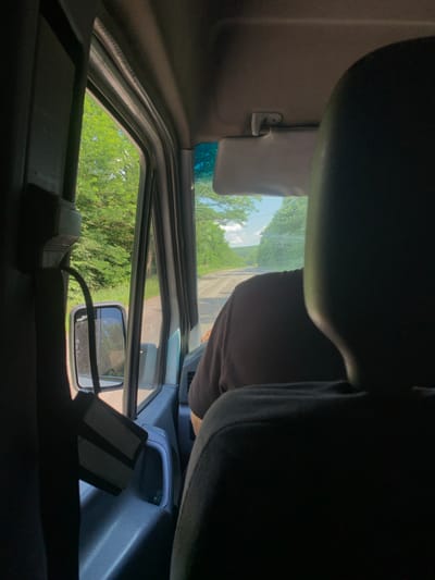 This image appears to be taken from inside a vehicle, likely a van or truck, looking out through the driver's side window and windshield. The perspective is unusual, as the camera seems to be positioned low and looking upwards and to the side, possibly from the driver's seat.
The interior of the vehicle is visible, including the dark headliner, a sun visor that is flipped down, and a portion of the dashboard. Through the window, we see a bright, sunny outdoor scene with lush green trees and a bright blue sky with some scattered clouds, suggesting it is daytime and the weather is fair. A road or path is visible in the distance through the windshield.
A side mirror is mounted on the driver's side door frame, reflecting the outdoor scenery. There is a dark-colored object with two lighter rectangular sections attached to the dashboard, possibly an electronic device or accessory. The overall impression is a snapshot of a journey through a rural or semi-rural area on a pleasant day. The image does not contain any visible text. The location context provided is Vashkivtsi, Ukraine.