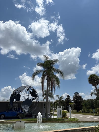 This is a vertically oriented, outdoor, daytime photograph taken in Bradenton, United States. The scene features a large, metallic globe sculpture with an open framework, depicting continents. In front of the globe, a blue SUV is parked. To the right of the globe, a water fountain is active, with jets of water spraying upwards into a shallow, rectangular pool. The background includes a building, a road, palm trees, and lush green foliage, suggesting a tropical or subtropical environment. The sky is a vibrant blue, dotted with fluffy white cumulus clouds, indicating fair weather. A speed limit sign indicating "10" is visible on a post, along with other signage that is partially obscured. The overall mood is bright and cheerful, enhanced by the sunny weather and the presence of water features.