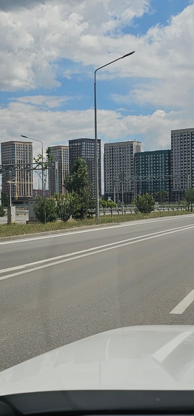 The image shows a street scene in the city of Овощной, Russia. In the background, there are several multi-story residential buildings. The buildings are modern in design, with numerous windows and balconies. In the foreground, there is a paved road with white lane markings. A white car is visible at the bottom of the frame, suggesting the photo was taken from inside a vehicle. There are streetlights and some vegetation along the side of the road. The sky is partly cloudy.