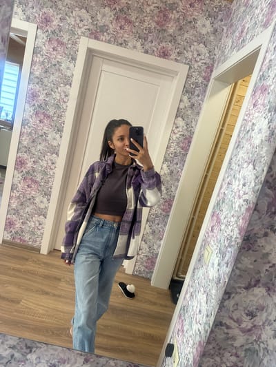 In a room with floral wallpaper and wooden floors, a young woman is taking a mirror selfie. She has dark hair pulled into a ponytail and is wearing a purple and white plaid shirt over a dark crop top and light-wash jeans. She is holding a black smartphone in her right hand. To her left, a white door is visible. A hallway with wooden walls is visible through an open doorway to her right. A white slipper with a heart-shaped pom-pom is on the floor near her feet. The scene appears to be indoors, possibly in a residential setting. The location is Oseyevo, Russia.