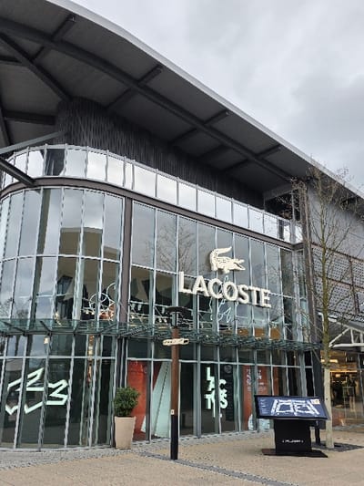 The image depicts the exterior of a retail store, identified as "LACOSTE," situated within the Zweibrücken Fashion Outlet in Zweibrücken, Germany. The building features a modern design with extensive glass curtain walls forming the lower two levels, including a prominent curved corner section on the left. The upper section and roof are constructed from dark gray, possibly metal, cladding with visible structural beams supporting an overhang.

The "LACOSTE" brand name, in white capital letters, along with its crocodile logo, is prominently displayed on the glass facade. Below this, and on the curved glass section to the left, additional white neon-style lettering or patterns are visible through the glass.

In the paved foreground, composed of light-colored interlocking pavers with dark linear accents, several objects are present. A tall, light-colored planter containing a small green bush stands near the entrance. To its right, a dark brown, multi-directional signpost with a black base and an integrated light fixture extends several arms. Further to the right, a black digital kiosk displays a blue and white map, likely a directory for the shopping complex. A leafless deciduous tree with thin branches is visible on the far right. The sky overhead is overcast and light gray. No individuals are present in the scene, and no specific actions or interactions are depicted.