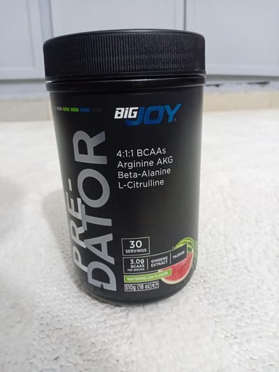 The image shows a black plastic container of "PRE-DATOR" pre-workout supplement by BIG JOY. The container is cylindrical with a ribbed black lid. The label features the brand name "BIG JOY" in blue, and the product name "PRE-DATOR" in large silver letters vertically oriented on the front. Below this, in smaller white text, are listed ingredients: "4:1:1 BCAAs", "Arginine AKG", "Beta-Alanine", and "L-Citrulline". Further down, it indicates "30 SERVINGS", "3.0g BCAAs PER SERVING", and "WATERMELON FLAVOR". A slice of watermelon is depicted on the lower right side of the label. The container is placed on a textured white surface, possibly a towel or fabric, in an indoor setting. The background is blurred and appears to be a wall with vertical elements. There are no people visible in the image. The setting is not identifiable as being in the city of Douala V, Cameroon.