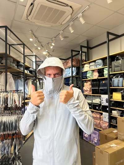 This image depicts a single individual inside a retail store located in Thành phố Kon Tum, Vietnam. The person, appearing male, is positioned centrally, facing the camera, wearing a light grey, full-zip jacket with an integrated hood and face covering that extends below the eyes. His eyes are visible and directed towards the viewer. Both hands are raised to chest level, with thumbs extended upwards in a gesture of approval.
The background shows two parallel retail shelving units with black metal frames and light wooden shelves. The left unit displays various clothing items, including lightweight jackets on hangers and smaller packaged items on a lower rack. The right unit showcases a diverse range of bags, including backpacks, tote bags, and handbags, in multiple colors such as pink, blue, green, black, and yellow. Cardboard boxes, some bearing the "TOKYOLIFE" label, are stacked on the floor in the lower right. The ceiling features white tiles, a prominent central cassette air conditioning unit, and a white track lighting system with multiple spotlights. A small security camera is also visible mounted on the track lighting.
