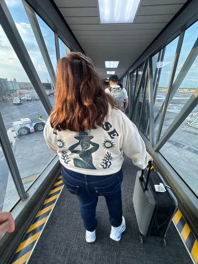 In Apodaca, Mexico, at an airport, a person with long brown hair and a white crewneck sweatshirt with a dark green graphic walks down a jet bridge, pulling a gray rolling suitcase. The sweatshirt has a graphic of a person wearing a hat with their hands on their head, a sun, and coral or seaweed. The person is wearing dark blue jeans and white and blue sneakers. Another person with a gray backpack is visible ahead, walking in the same direction. The jet bridge has large glass windows on the sides, offering a view of the tarmac where a tug vehicle with "FBT 74" and "M" visible on its side is parked. The sky is partly cloudy and appears to be late afternoon or early evening.